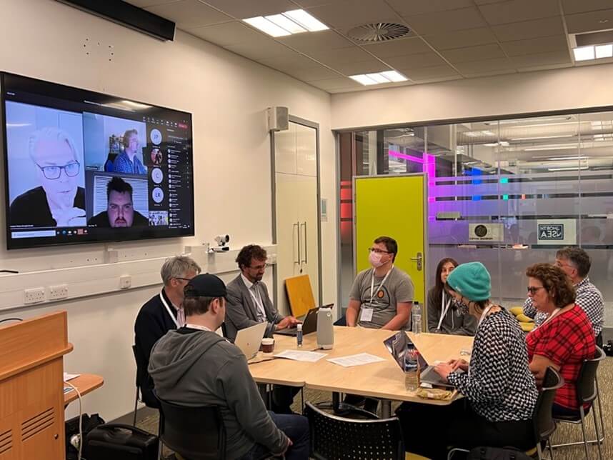 The image shows a group of delegates around a table sitting next to a wall with a video screen where other delegates have joined the conference virtually. In front of the delegates in the room there is a lectern and behind them a glass wall with a door.